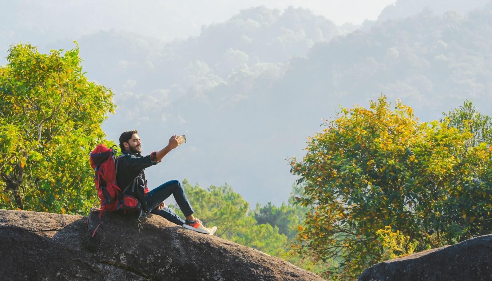 Asian man traveler using mobile phone taking selfie while solo travel on top mountain