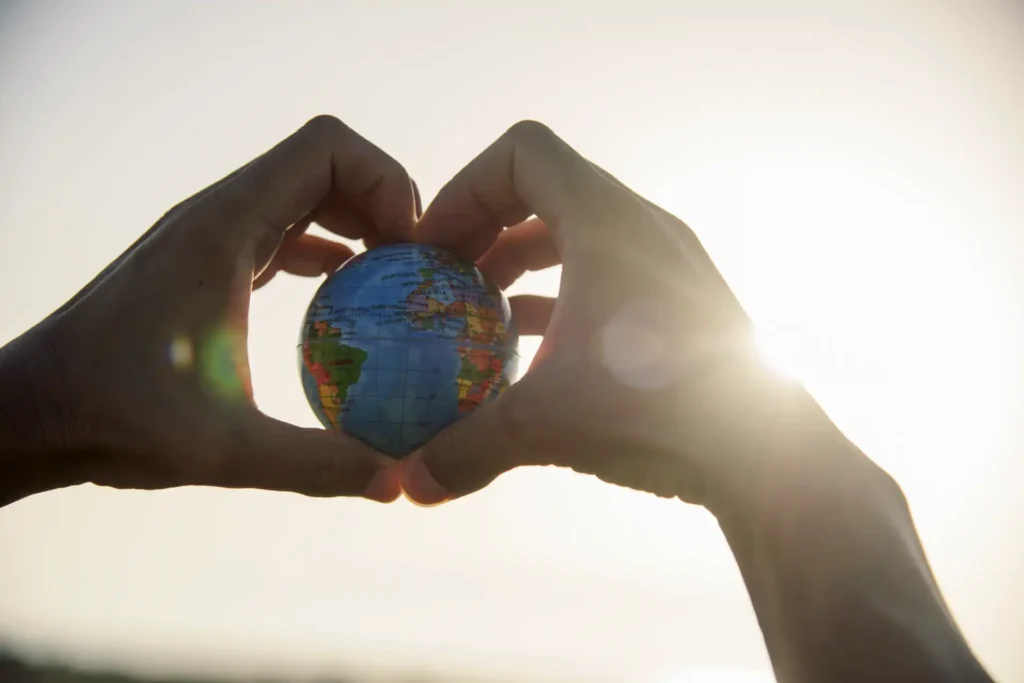 young caucasian man with a world globe in his hands forming a heart against the sky and a ray of sun showing up on the right