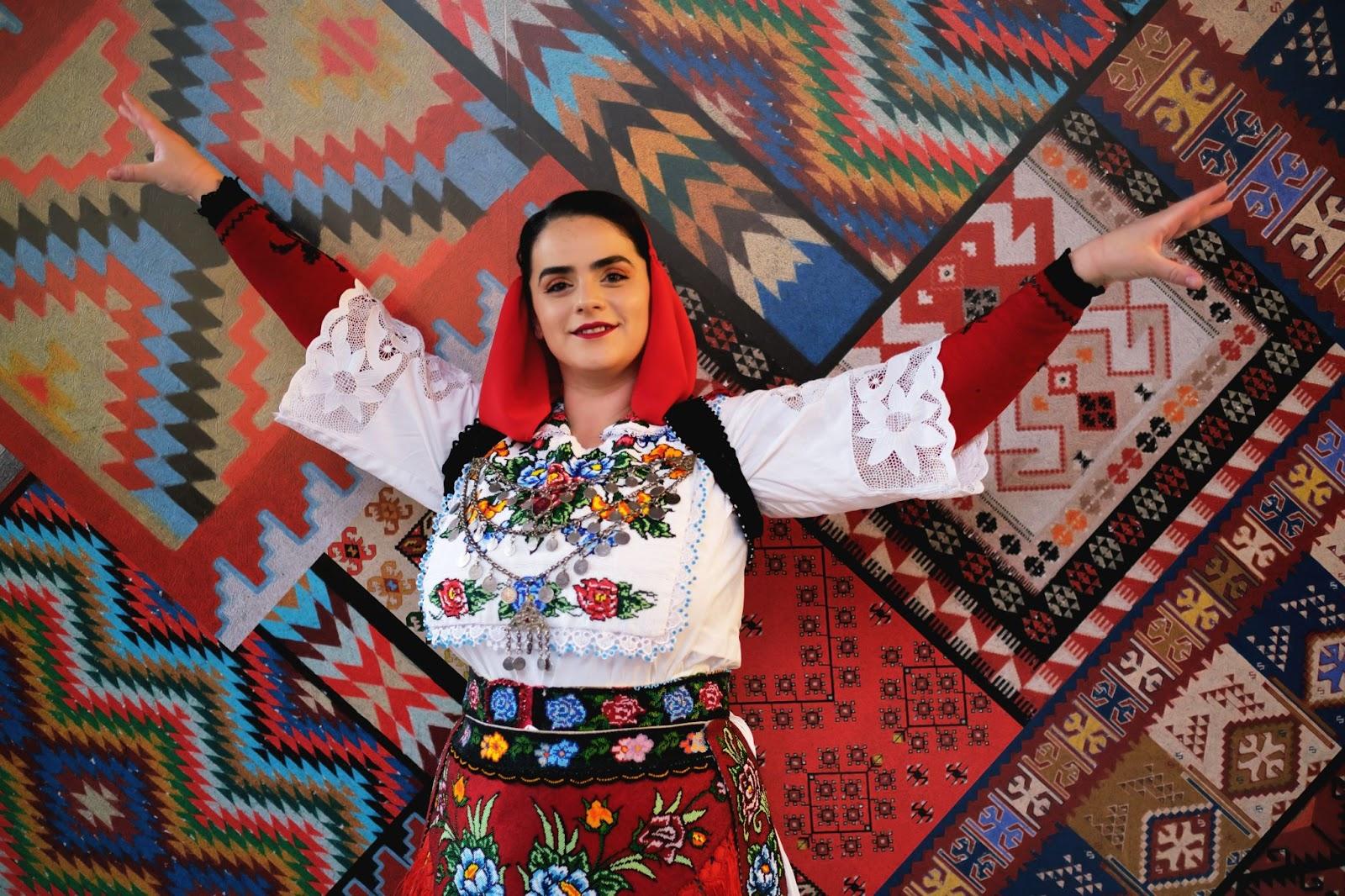 A woman in traditional Albanian clothing poses alone against a backdrop with traditional Albanian patterns