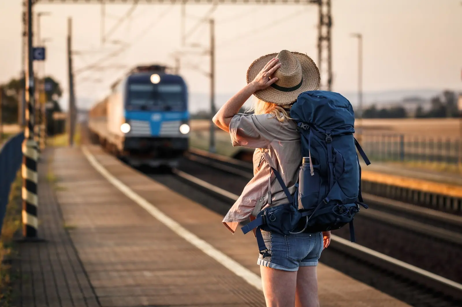 Travel by train. Woman backpacker with hat standing at railroad station and looking to arriving train