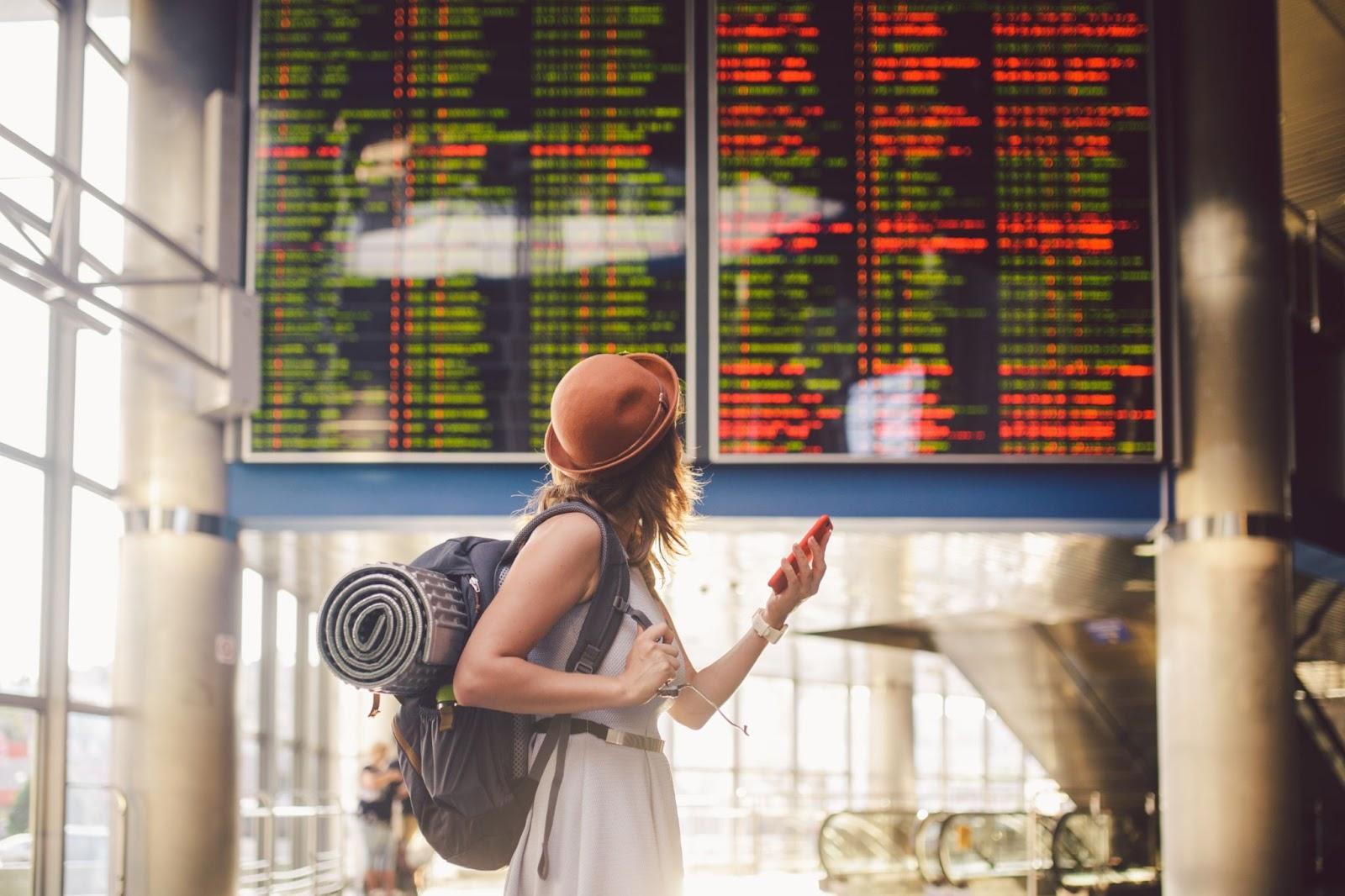 Theme travel and tranosport. Beautiful young caucasian woman in dress and backpack standing inside train station or terminal looking at a schedule holding a red phone, uses communication technology.