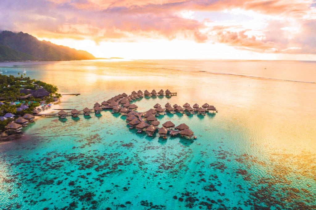 Luxury travel vacation aerial of overwater bungalows resort in coral reef lagoon ocean by beach. View from above at sunset of paradise getaway Moorea, French Polynesia, Tahiti, South Pacific Ocean.