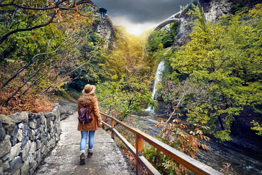 Tourist Woman in Hat going to waterfall in Botanical Garden with autumn trees at overcast cloudy sky in Tbilisi, Georgia